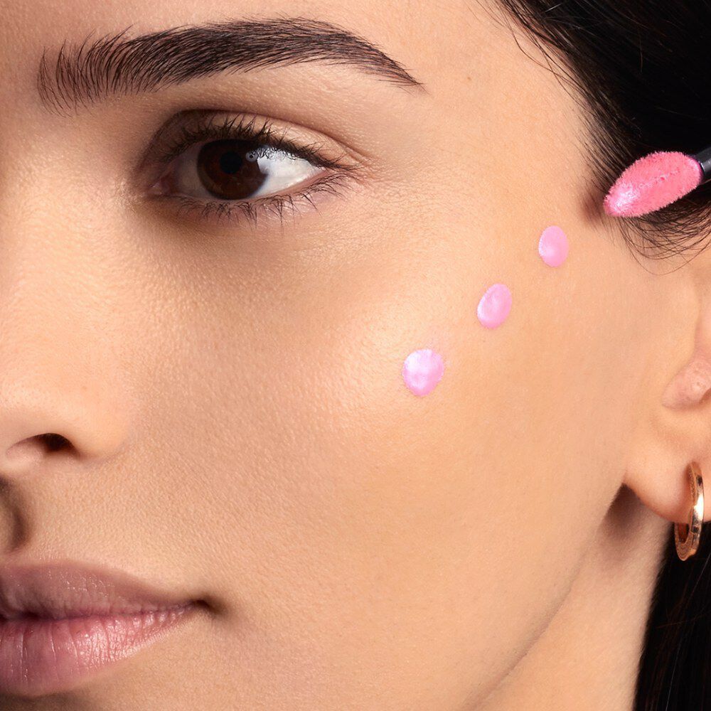 La joue d'une femme avec cinq points distincts de produit liquide rose clair, probablement un blush ou un illuminateur, deposes le long de la pommette, prets a etre estompes.