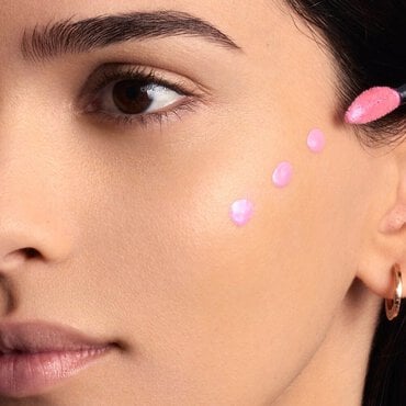 La joue d'une femme avec cinq points distincts de produit liquide rose clair, probablement un blush ou un illuminateur, deposes le long de la pommette, prets a etre estompes.