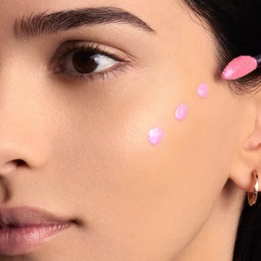 A woman's cheek is shown with five distinct dots of light pink liquid product, likely a blush or highlighter, dotted along her cheekbone area, ready for blending.