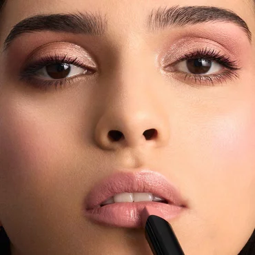 A close-up of a woman's face as she applies a light pink or nude lipstick to her upper lip.
