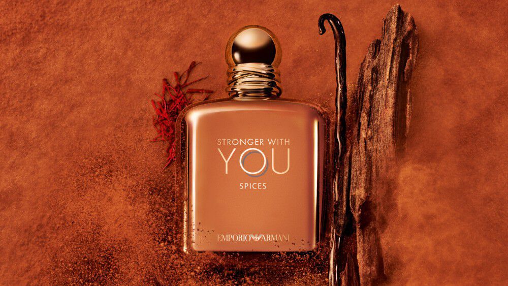 ARMANI STRONGER WITH YOU SPICES bottle with gold cap, placed among vanilla beans and saffron on an orange textured background, highlighting the fragrance’s spicy, gourmand notes.