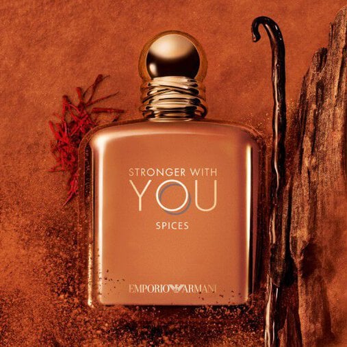 ARMANI STRONGER WITH YOU SPICES bottle with gold cap, placed among vanilla beans and saffron on an orange textured background, highlighting the fragrance’s spicy, gourmand notes.