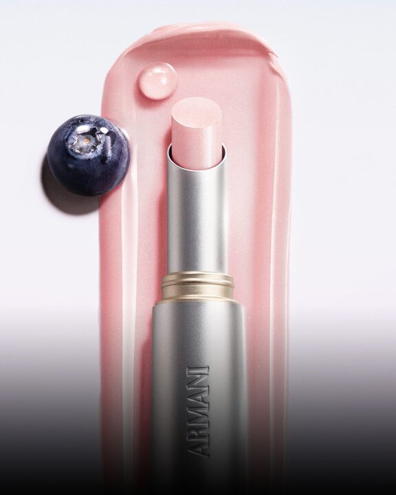 Armani Prisma Flash Petal Flash lipstick, captured in use, featuring its soft pink hue and elegant metallic packaging. Next to a blueberry fruit.