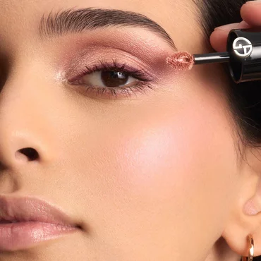 A woman applies shimmery liquid eyeshadow in a rosy bronze shade to her eyelid using the product's applicator wand.