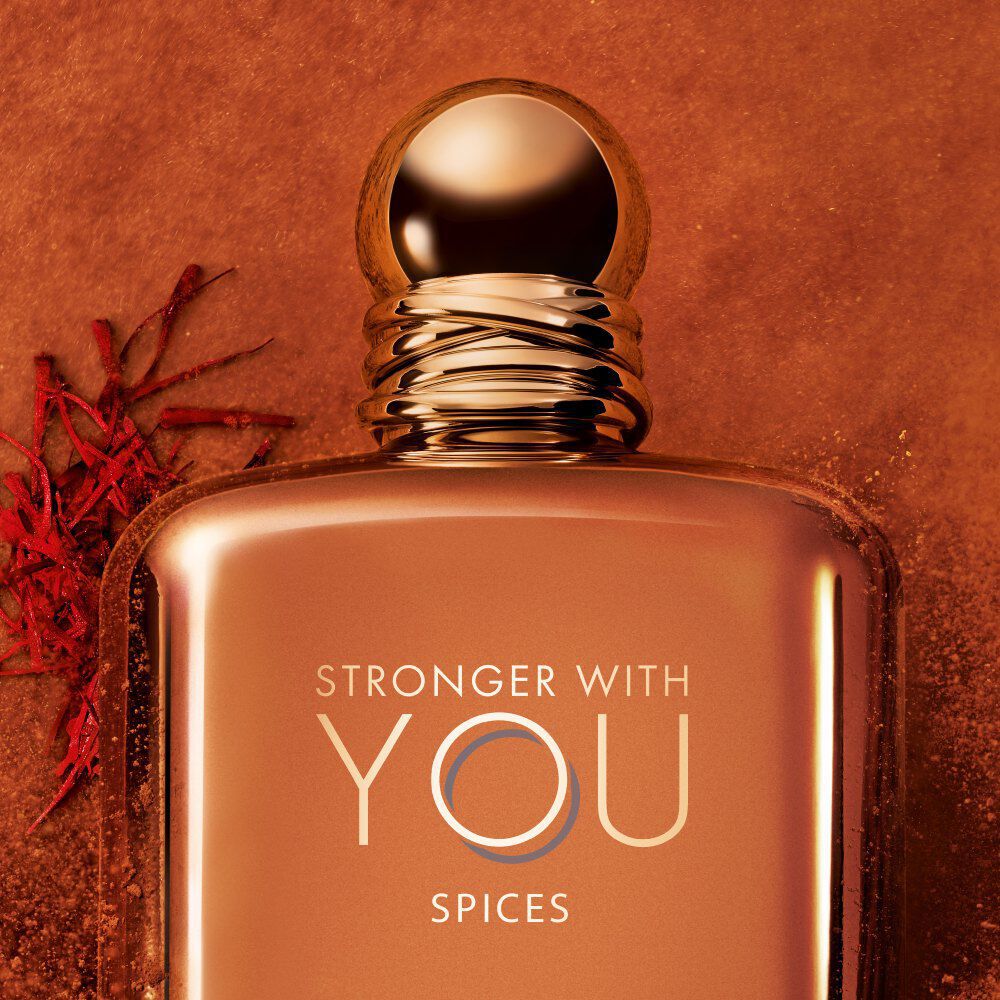 Close-up of ARMANI STRONGER WITH YOU SPICES bottle with gold cap, set against an orange background with saffron and spice accents, showcasing the luxurious and intense scent.