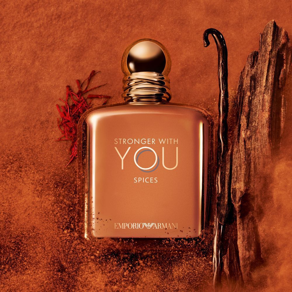 ARMANI STRONGER WITH YOU SPICES bottle with gold cap, placed among vanilla beans and saffron on an orange textured background, highlighting the fragrance’s spicy, gourmand notes.