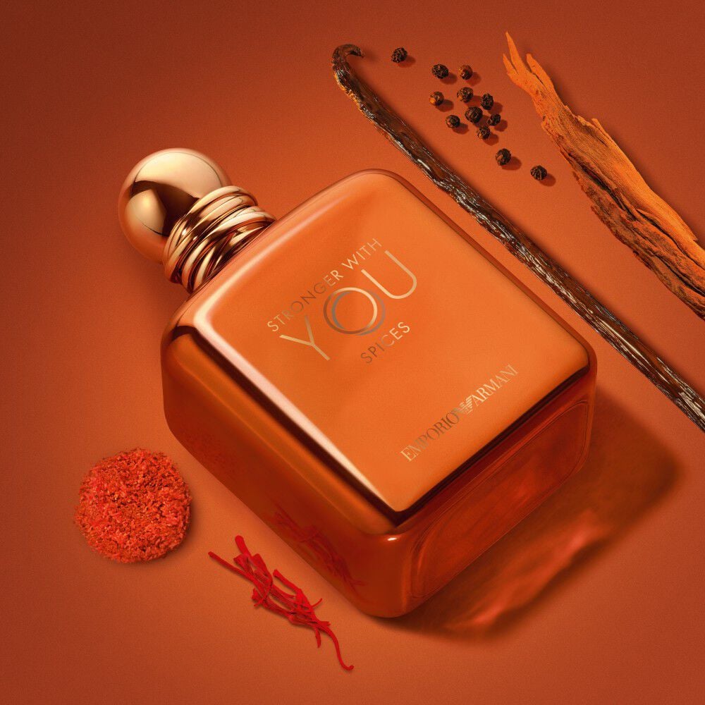 ARMANI STRONGER WITH YOU SPICES bottle with gold cap, surrounded by vanilla bean, saffron, and spices on a warm orange background, highlighting the fragrance’s bold and spicy character.
