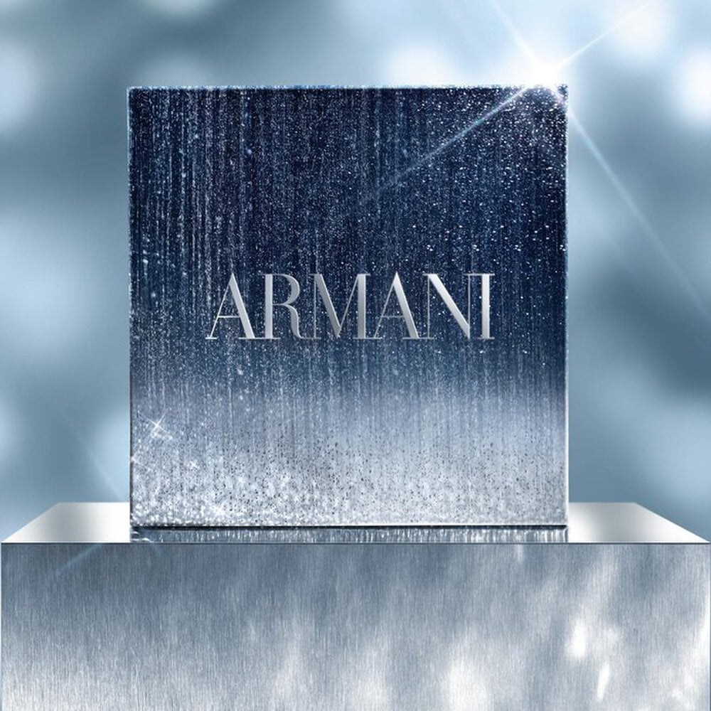 A square Armani GIFT SET in a shimmering silver and blue gradient box, displayed on a reflective pedestal with a soft, sparkling light background.