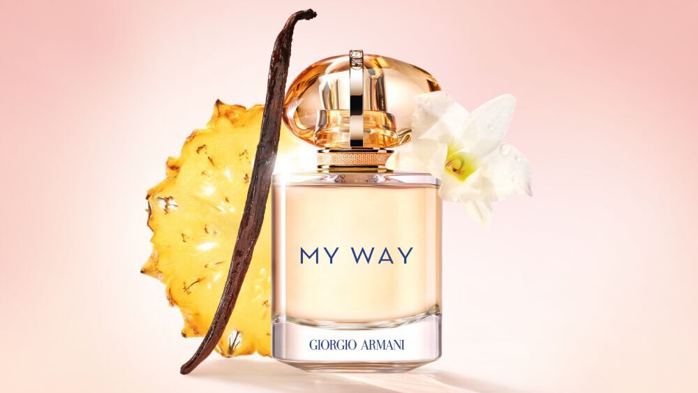 Armani My Way Sunny Vanilla perfume bottle displayed with a slice of pineapple, a vanilla bean, and tuberose flower, set against a soft pink background to highlight its fresh and natural ingredients.