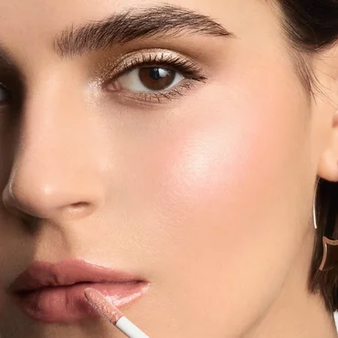 A close-up of a woman's face as she applies a light pink or nude lipstick to her upper lip.