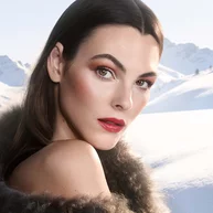 A model embodies winter elegance — bold rouge lip, luminous sculpted skin, intensely defined eye — set against glacial alpine peaks. Armani beauty Holiday 2026. Square.