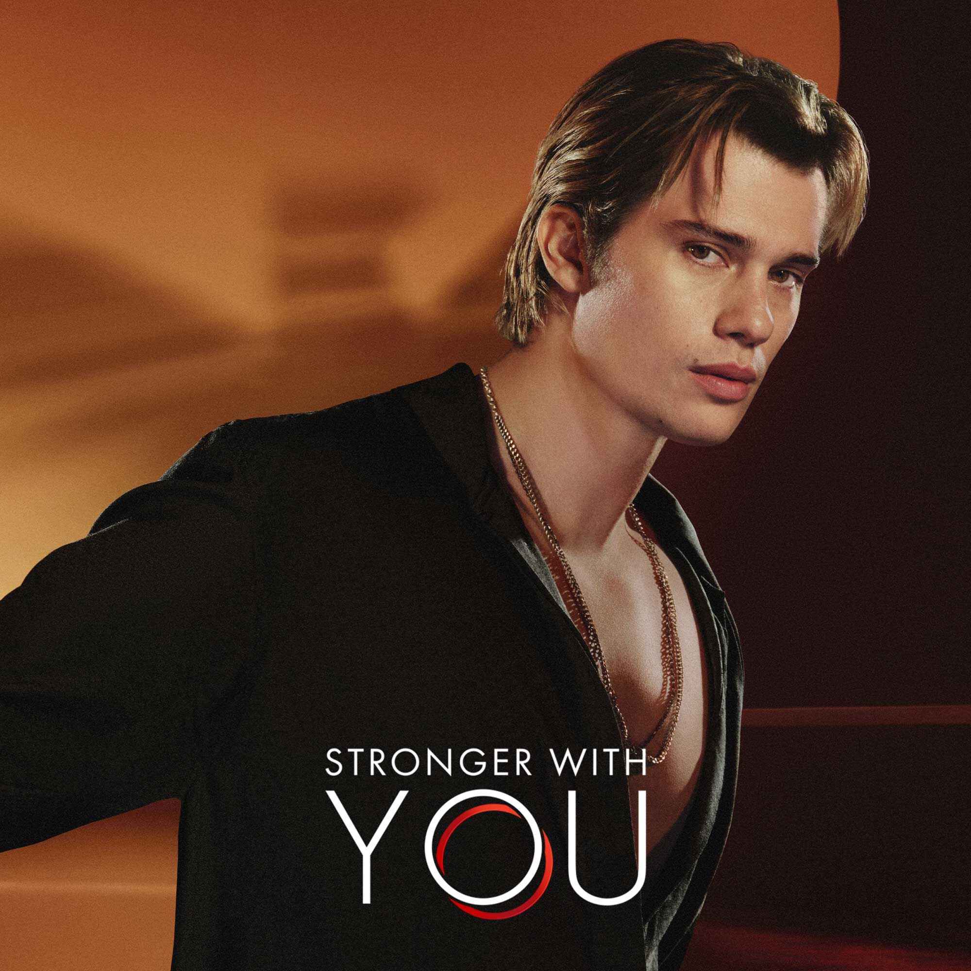 Nicholas Galitzine in a stylish Armani campaign for "Stronger With You" fragrance, wearing a black open shirt against a warm, elegant backdrop.