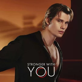 Nicholas Galitzine in a stylish Armani campaign for "Stronger With You" fragrance, wearing a black open shirt against a warm, elegant backdrop.