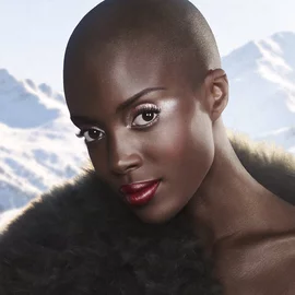 A model embodies winter elegance — Deep skin radiates an even, luminous glow. Eyes framed by full lashes and sculpted brows. A deep lacquered red lip as the defining statement. Snow-capped peaks behind. 