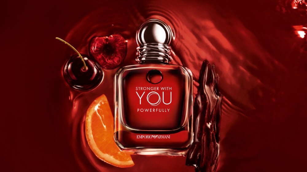 Emporio Armani Stronger With You Powerfully bottle with cherries, orange slice, and wood, on a vibrant red backdrop.